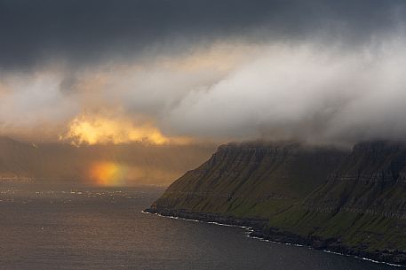 Beauttiful light and rainbow through the dense clouds of the coast, Faeroe islands, Denmark, Europe